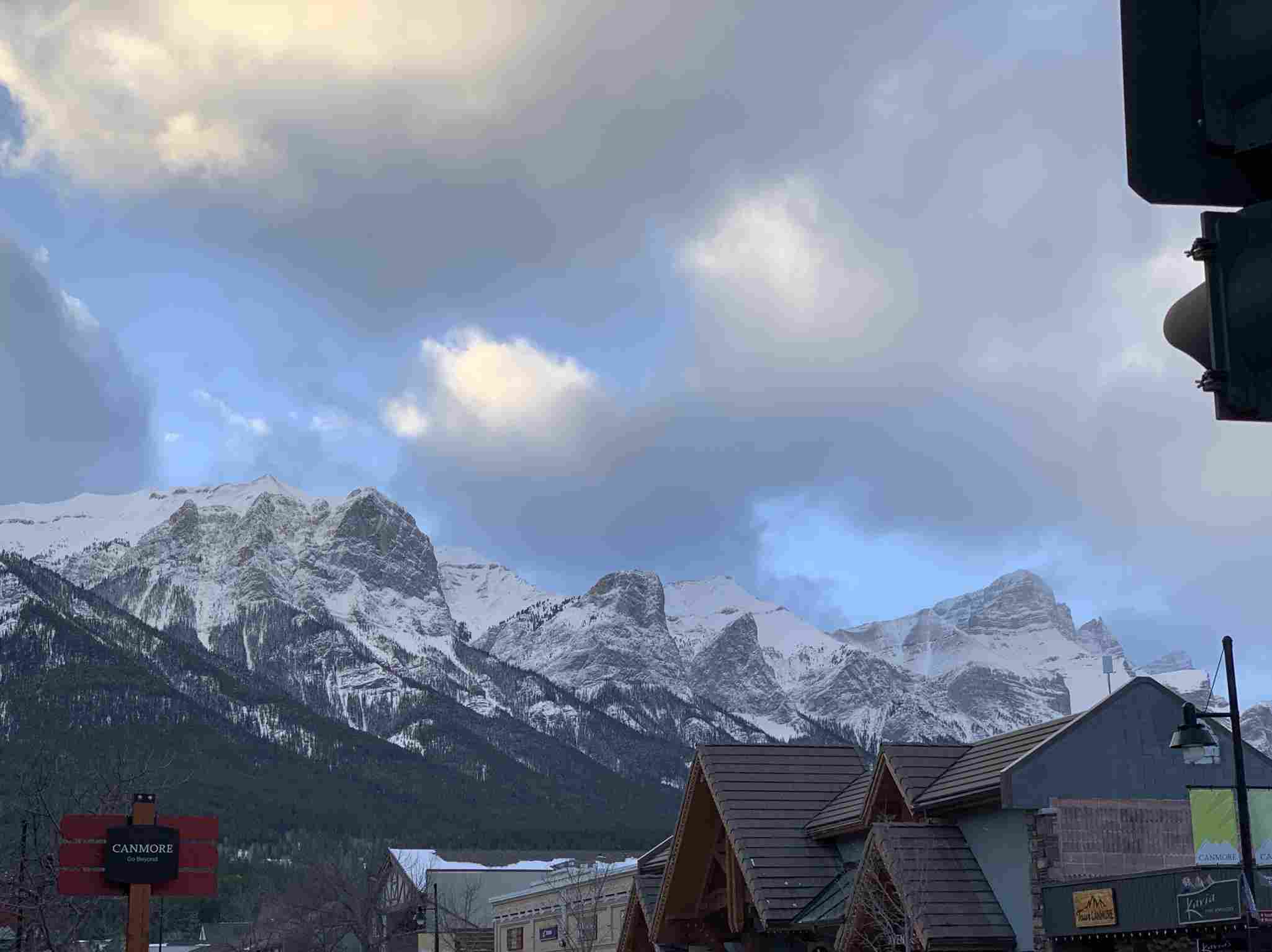 The Rocky Mountains as seen from Canmore, Alberta.  The sky is partly cloudy, with blue showing between.  The mountains are covered in snow, and the roofs of houses are visible along the bottom of the image.