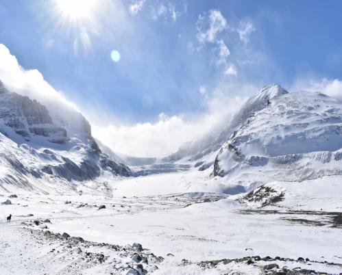 A snowy scene in the mountains.  It is a bright, sunny day with blue skies and clouds, and the wind is picking up snow.