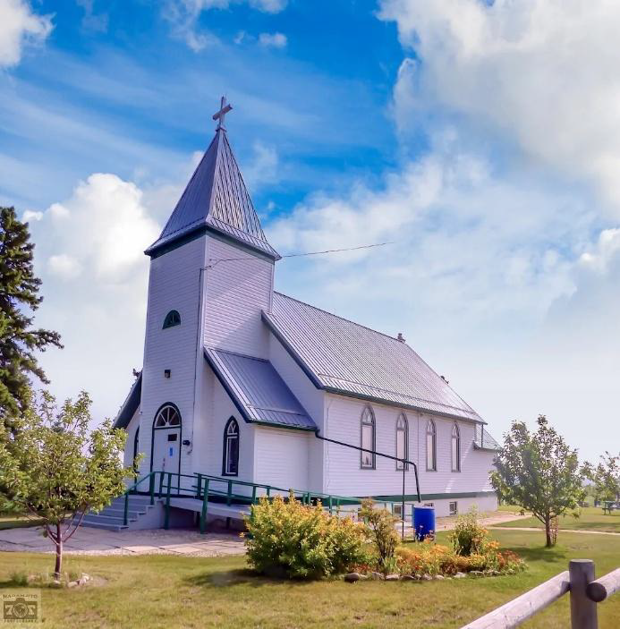 An image of Mewassin United Church.  The front and side of the building are visible, the sky is blue and partly cloudy and the trees are leafy green